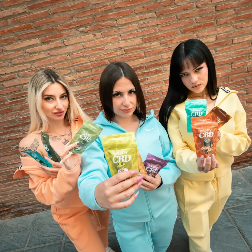 Tres mujeres con ropa colorida sosteniendo productos de CBD frente a una pared de ladrillos.